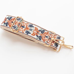 May include: A white fabric strap with a gold metal buckle and hook. The strap has a repeating pattern of orange, blue, and green floral designs.