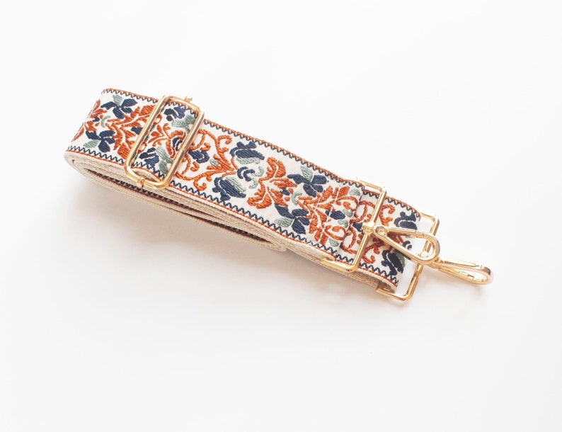 May include: A white fabric strap with a gold metal buckle and hook. The strap has a repeating pattern of orange, blue, and green floral designs.