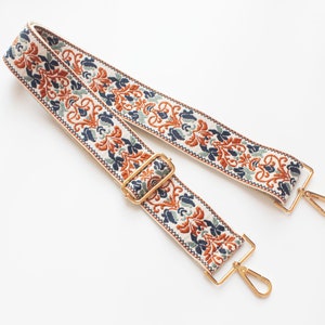 May include: A white fabric strap with a blue, green, and orange floral pattern. The strap has gold metal clasps on each end.