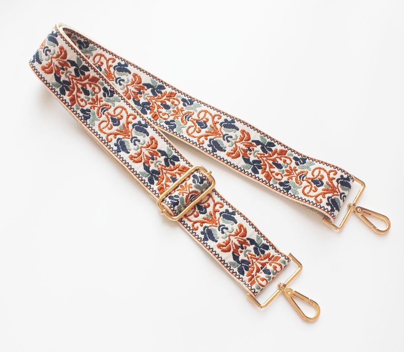 May include: A white fabric strap with a blue, green, and orange floral pattern. The strap has gold metal clasps on each end.