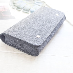 May include: A rectangular gray felt case with rounded edges and a flap closure. The flap is secured by two silver-toned metal snap buttons positioned on the front surface. The case has a thick, textured construction and a simple, functional design.