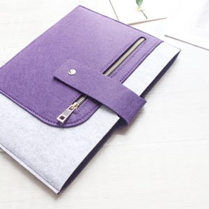 May include: A purple and grey felt laptop sleeve. It features a silver zip closure and a strap with a snap. The sleeve is rectangular and designed to protect a laptop or tablet. The zip and snap are silver.