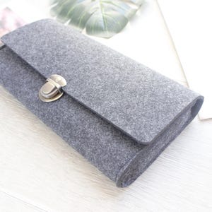 Può includere: Un portafoglio rettangolare in feltro grigio scuro con una superficie in tessuto testurizzato e una chiusura a girello in metallo color bronzo sulla patta anteriore. Il portafoglio ha una forma allungata e strutturata con bordi arrotondati.