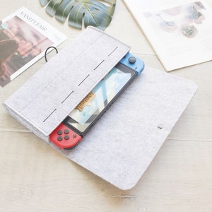 Könnte beinhalten: Eine hellgraue Filzhülle mit schwarzem Gummizugverschluss, die ein blaues und rotes Handheld-Spielgerät enthält. Die Hülle ist geöffnet und zeigt den Bildschirm und die Controller. Die Hülle liegt auf einer hellen Holzoberfläche.