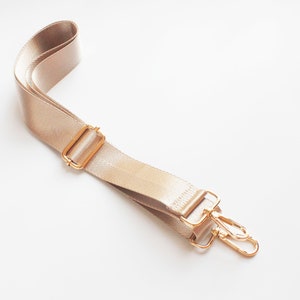 May include: A beige adjustable strap with gold metal hardware. The strap has a buckle and two swivel hooks.