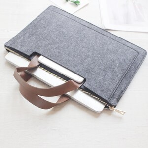 May include: A gray felt laptop sleeve with a brown leather handle and a gold zipper. A laptop and a smartphone are partially visible inside the case. The case is designed to protect and carry electronic devices.