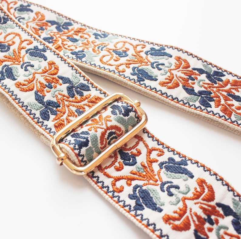 May include: A white fabric strap with a gold metal buckle. The strap has a colorful embroidered pattern with orange, blue, and green floral designs.