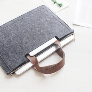 May include: A gray felt laptop sleeve with a brown leather handle. The sleeve has a zipper closure and is designed to hold a laptop and a phone. The laptop sleeve is on a white surface.