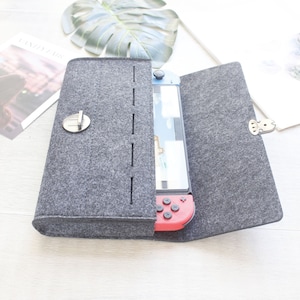 May include: A gray felt case with a zipper closure and a built-in pocket for a Nintendo Switch console. The case is open and the console is visible inside.