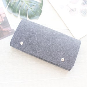 Pode incluir: Uma clutch cinza de feltro com dois fechos de press&atilde;o prateados. A bolsa retangular est&aacute; dobrada e repousa sobre uma superf&iacute;cie branca, com uma revista e uma grande folha verde ao fundo.