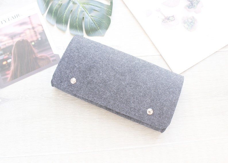Pode incluir: Uma clutch cinza de feltro com dois fechos de press&atilde;o prateados. A bolsa retangular est&aacute; dobrada e repousa sobre uma superf&iacute;cie branca, com uma revista e uma grande folha verde ao fundo.