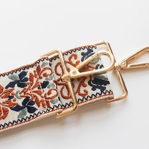 May include: A close-up of a cream-colored fabric strap with a gold-toned metal clasp and hook. The strap has a colorful floral pattern in shades of orange, blue, and green.