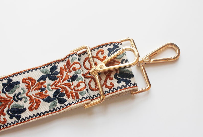 May include: A close-up of a cream-colored fabric strap with a gold-toned metal clasp and hook. The strap has a colorful floral pattern in shades of orange, blue, and green.