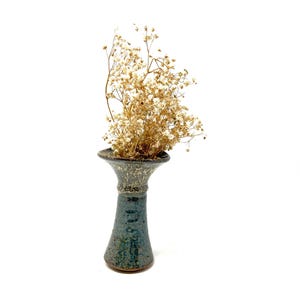 May include: A tall, ceramic vase with a flared top and a dark teal glaze. The vase is filled with dried, light brown flowers. The vase has a textured, speckled design around the top. The vase is a decorative item.