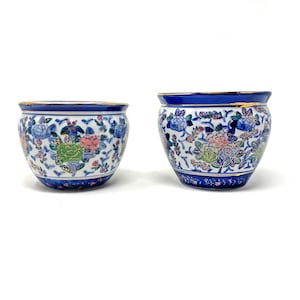 May include: Two ceramic bowls featuring a white base with a blue, green, and pink floral pattern. Each bowl has a deep blue rim and base, accented with a thin gold-colored trim along the top edge. The bowls have a rounded, bulbous shape.