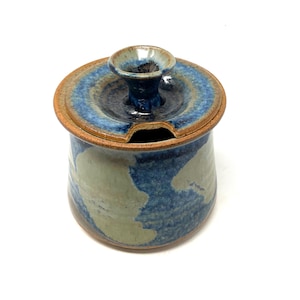 May include: A handmade ceramic jar with a lid. The jar is cylindrical with a blue and gray mottled glaze. The lid is circular with a small handle and a slot for a spoon. The rim is a natural brown color.