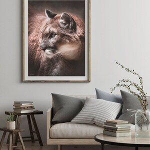 May include: A framed photograph of a brown and white cougar with green eyes looking to the left. The cougar is in a dark brown setting. The frame is made of wood with a light brown finish.
