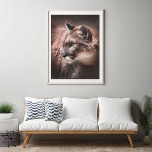 May include: A close-up portrait of a brown and tan cougar with green eyes. The cougar is looking to the left of the frame. The background is a dark brown color.