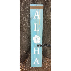 May include: A wooden sign painted light blue with the word "ALOHA" spelled vertically in white letters. A white hibiscus flower is painted between the "L" and "H". The sign is attached to a tree with twine.