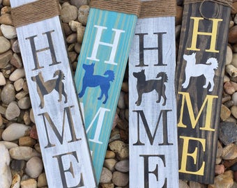 May include: Three wooden signs with the word "HOME" spelled out in white, black, and yellow paint. Each sign features a silhouette of a dog in a different color. The signs are tied with twine.