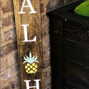 May include: A wooden sign with the word "Aloha" painted in white letters. A yellow pineapple with green leaves is painted between the "L" and the "H". The sign is attached to a brick wall with a piece of twine. The text "Fisch Tale" is written in cursive at the bottom of the sign.