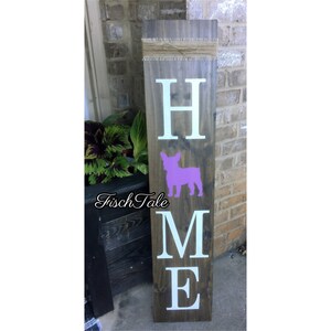 Op de afbeelding: Een houten bord met het woord "HOME" in witte letters. Tussen de "H" en de "M" staat een paarse silhouet van een Franse Bulldog. Het bord is op een donkerbruine houten achtergrond.