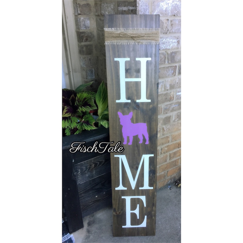 Op de afbeelding: Een houten bord met het woord "HOME" in witte letters. Tussen de "H" en de "M" staat een paarse silhouet van een Franse Bulldog. Het bord is op een donkerbruine houten achtergrond.