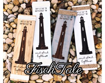 May include: Four wooden signs with a lighthouse silhouette and the coordinates 30°11'16" N 88°03'02" W. The signs are painted in different colors: white, light blue, brown, and dark brown. The text "sand island lighthouse" is written on each sign. The signs are arranged in a row on a bed of small, round stones. The text "Fisch Tale" is written in a decorative font below the signs.
