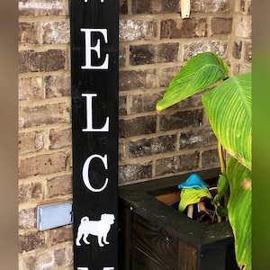 May include: Black wooden welcome sign with white letters spelling "WELCOME" and a white silhouette of a pug dog. The sign has a brown burlap bow with a white flower on top. The sign is leaning against a brick wall with a wooden planter box below it.