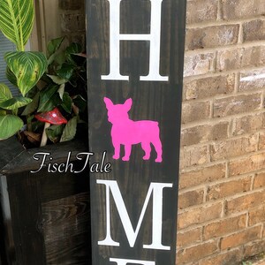 Op de afbeelding: Een houten bord met het woord "HOME" in witte letters. Tussen de "H" en de "M" staat een roze silhouet van een Franse bulldog. Het bord is versierd met een bruin jute lint met een strik.