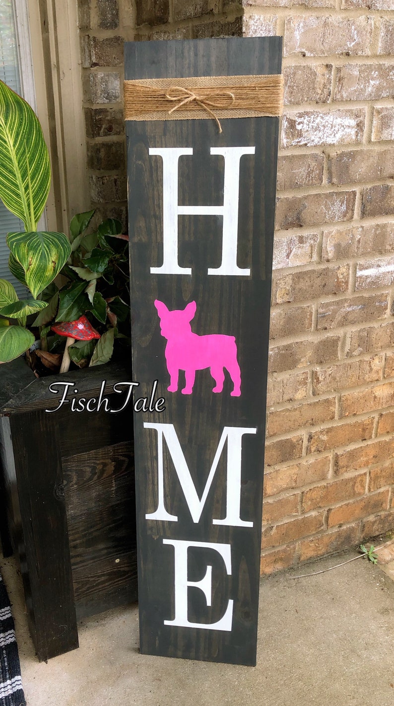 Op de afbeelding: Een houten bord met het woord "HOME" in witte letters. Tussen de "H" en de "M" staat een roze silhouet van een Franse bulldog. Het bord is versierd met een bruin jute lint met een strik.