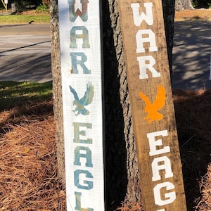 May include: Two wooden signs with the words "WAR EAGLE" painted on them. The signs are painted in different colors, one is white with blue and green accents and the other is brown with white letters. The signs are leaning against a tree trunk.