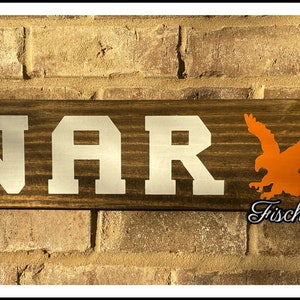 May include: A wooden sign with the word "WAR" in white letters and an orange silhouette of an eagle. The sign is hanging on a brick wall and has rope accents.