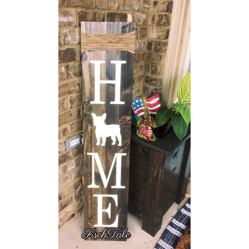 Op de afbeelding: Een houten bord met het woord "HOME" in witte letters. Tussen de "H" en de "M" staat een wit silhouet van een Franse bulldog. Het bord is gemonteerd op een bruine houten plank en heeft bovenaan een bruine touw strik.