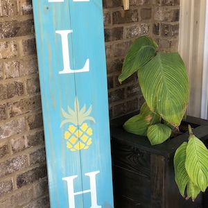 May include: A blue wooden sign with the word "Aloha" painted in white letters. A yellow pineapple graphic is between the "L" and the "H". The sign is decorated with a pink flower and twine.