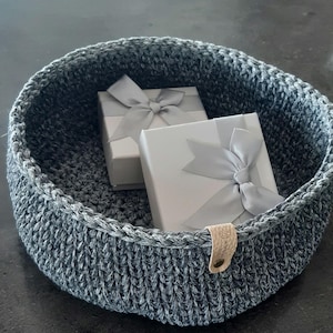 Può includere: Un cesto in crochet grigio con un manico in pelle, contenente due scatole regalo bianche con fiocchi argentati.