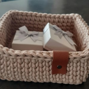 May include: A beige crocheted basket with a brown leather handle. Two white gift boxes with grey bows are inside the basket.