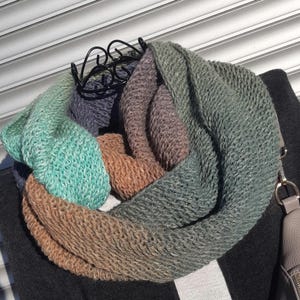 Pode incluir: Uma echarpe infinita de malha multicolorida com tons de verde, marrom e roxo. A echarpe é feita de um fio macio e texturizado e é perfeita para se manter aquecido no inverno.