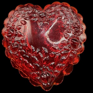 Può includere: Un piatto a forma di cuore in vetro rosso rubino con bordo smerlato. Il piatto è decorato con rose e foglie in rilievo. Il vetro ha una finitura lucida ed è posto su uno sfondo nero.