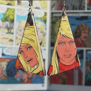 May include: A pair of triangular earrings with a comic book character design. The earrings feature a woman with blonde hair and a red and white outfit. The earrings are made of a hard material and have a glossy finish.