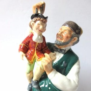 Puede incluir: Figura de cerámica que representa a un hombre sosteniendo a un niño. El hombre viste un chaleco verde, camisa blanca y pantalón marrón. El niño lleva una chaqueta roja y pantalones verdes. La figura tiene un aspecto detallado y pintado a mano.