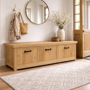 May include: A light oak entryway bench with three drawers and black handles. A round mirror hangs above the bench, with a coat and bag hanging on the wall. A patterned rug and vase of flowers complete the welcoming space.