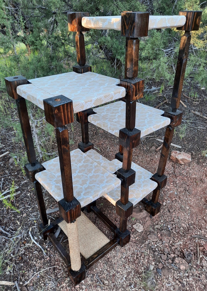 May include: A multi-level cat tree constructed from dark stained wooden posts and square frames. It features four platforms covered in a light beige fabric with a white geometric pattern and a base section wrapped in natural tan sisal rope.