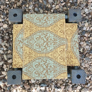 May include: A small, rectangular footstool featuring a fabric top with a repeating damask pattern in shades of mustard yellow and light teal. The stool is supported by four dark gray, square wooden legs, each with a circular hole in the center.
