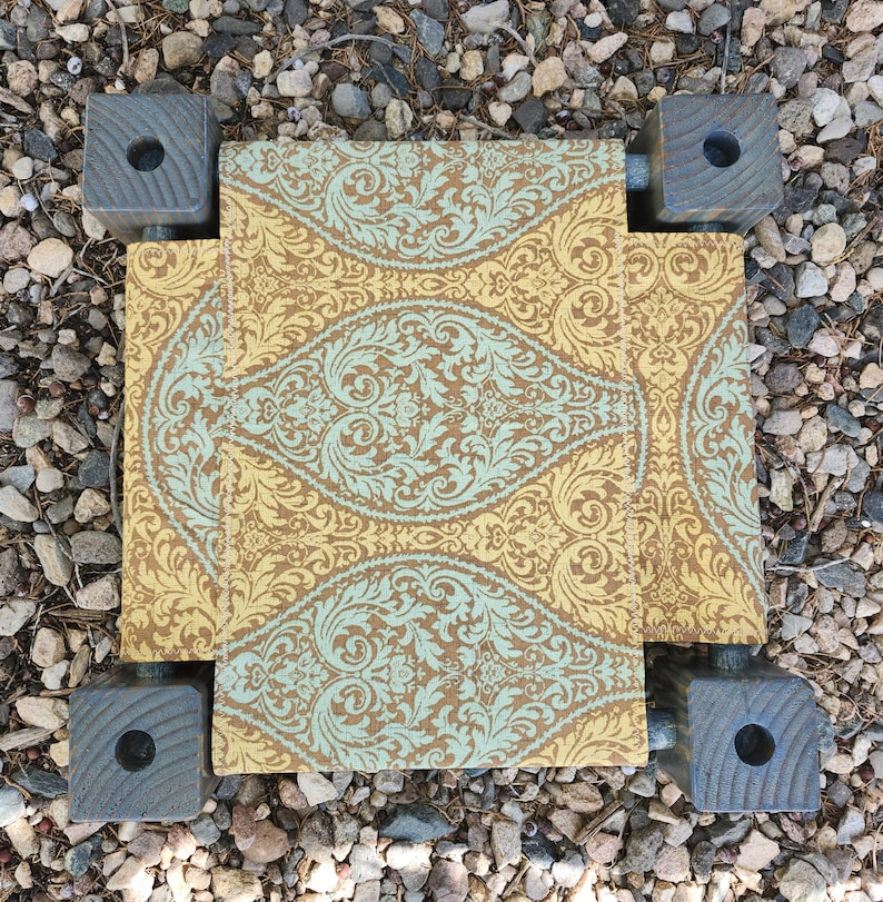 May include: A small, rectangular footstool featuring a fabric top with a repeating damask pattern in shades of mustard yellow and light teal. The stool is supported by four dark gray, square wooden legs, each with a circular hole in the center.