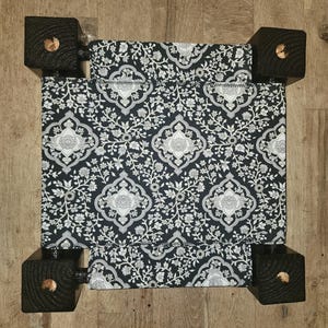 May include: A small pet bed featuring a black fabric cover with a white floral and damask-style pattern. The bed is supported by four square, dark-stained wooden legs at each corner, providing a raised structure for the fabric sleeping surface.