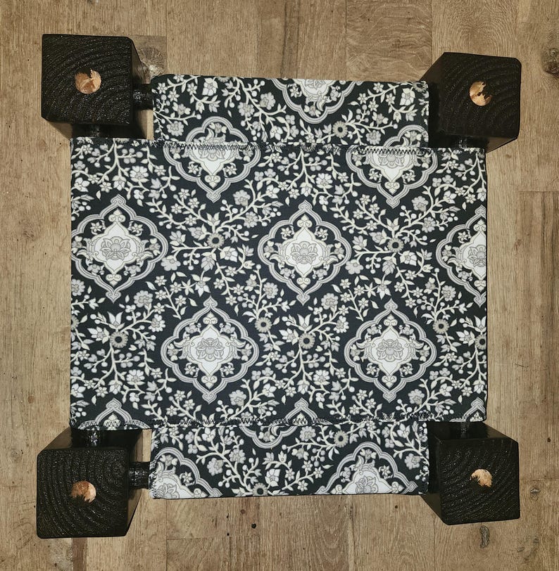 May include: A small pet bed featuring a black fabric cover with a white floral and damask-style pattern. The bed is supported by four square, dark-stained wooden legs at each corner, providing a raised structure for the fabric sleeping surface.