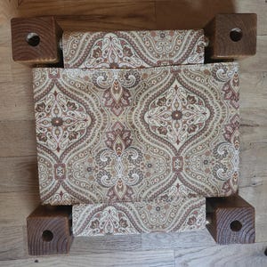 May include: A square footstool featuring four dark brown wooden legs with circular cutouts at the corners and a central seat upholstered in a fabric with a repeating tan, brown, and cream paisley pattern. The design shows a symmetrical layout.