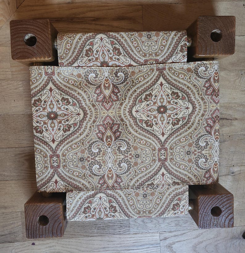 May include: A square footstool featuring four dark brown wooden legs with circular cutouts at the corners and a central seat upholstered in a fabric with a repeating tan, brown, and cream paisley pattern. The design shows a symmetrical layout.