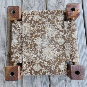 May include: A square footstool featuring a brown fabric top with a cream-colored floral damask pattern. The stool is supported by four thick, dark brown wooden legs, each with a circular hole drilled through the center of the top surface.